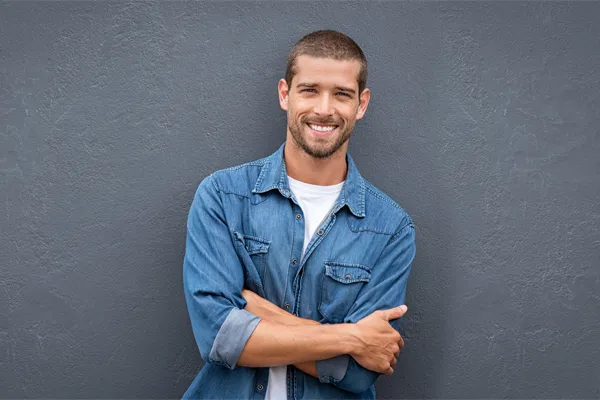 Hormones-for-Men-Doctor A man in a denim shirt stands smiling against a gray-blue wall, pleased with his testosterone hormone therapy from David Garcia, D.O. in Nashville Metro.