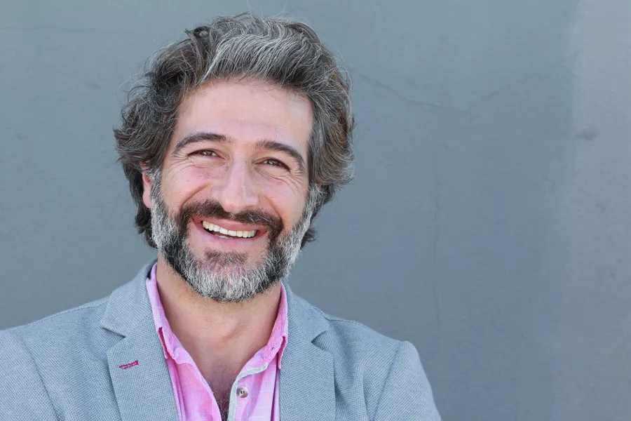 Hypogonadism-Clinic A smiling man with salt and pepper hair and beard wearing a pink shirt and gray blazer stands agains a gray wall. Get hypogonadism treatment from David Garcia, D.O. in Nashville Metro.