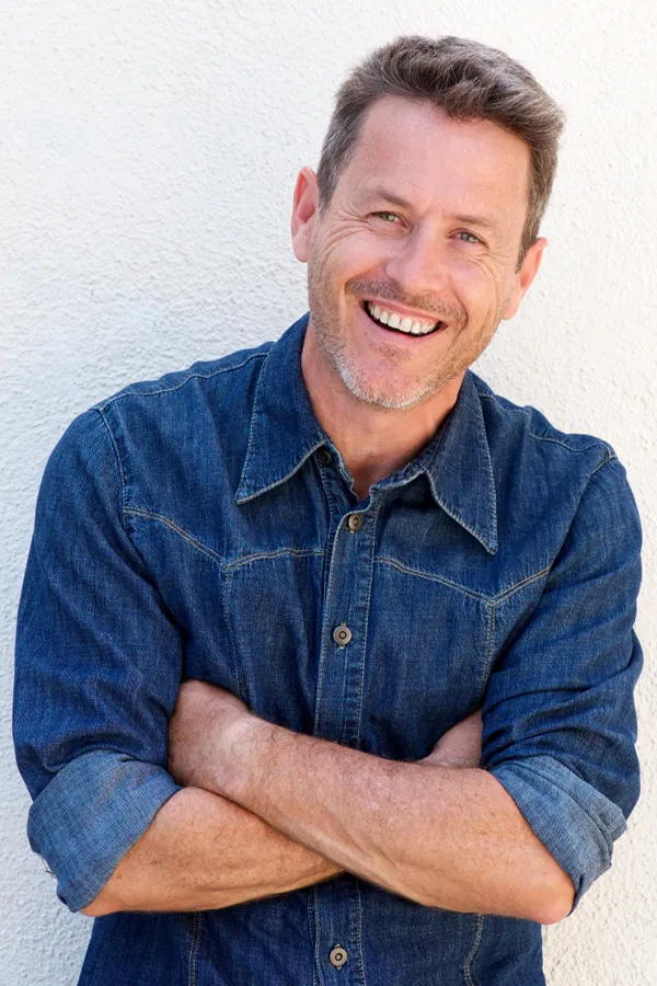 Ipamorelin-Peptide-Treatment A healthy middle-aged man in a denim shirt leans against a white wall smiling. Get Ipamorelin peptide treatment from David Garcia, D.O. in Nashville Metro.