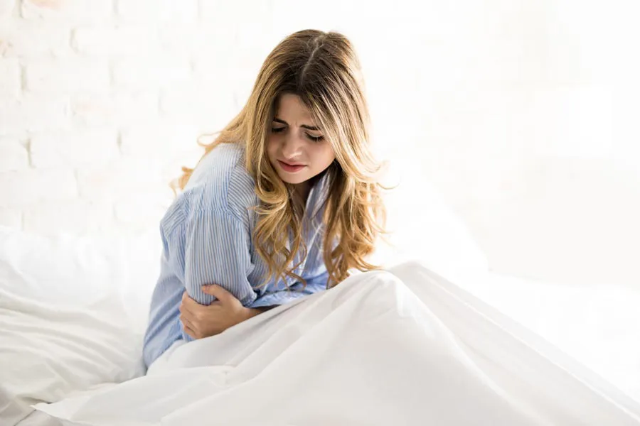 PMS-Therapy A woman with long blonde hair sits in her bed holding her stomach in pain, considering getting care for PMS from David Garcia, D.O. in Nashville Metro.