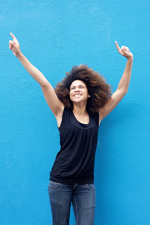 PMS-Treatment A woman in a dark blue tank top standing in front of a bright blue wall, raising her arms in celebration of relief from PMS from David Garcia, D.O. in Nashville Metro.