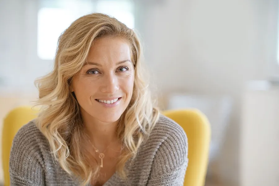 Perimenopause-Doctor A blonde woman wearing a a gray sweater sits inside in a yellow chair, smiling and happy with her perimenopause treatment from David Garcia, D.O. in Nashville Metro.