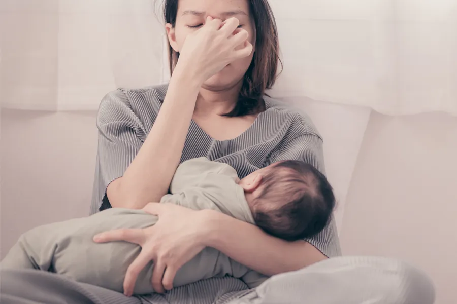 Postpartum-Hormone-Imbalance-Clinic A woman nursing an infant on couch rubs her eyes with exhaustion. Get treatment for postpartum hormone imbalance from David Garcia, D.O. in Nashville Metro.