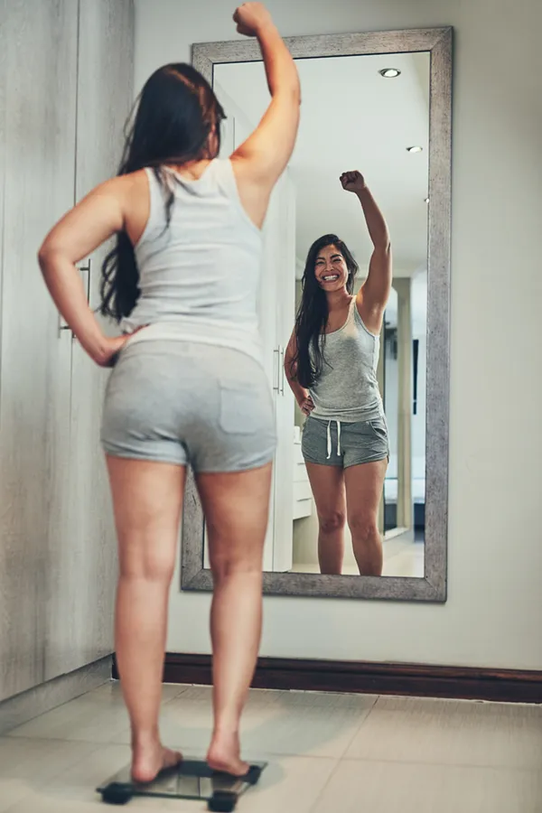 Tirzepatide-Treatment A woman in workout clothes stands on a scale looking in the mirror and pumping her hands in celebration of her successful Tirzepatide treatment from David Garcia, D.O. in Nashville Metro.